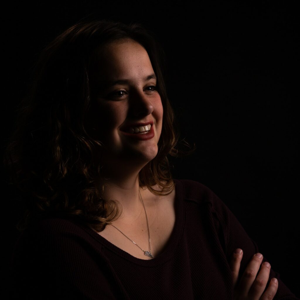 A girl with medium brow curly hair, and fair features stands against a dark background with her arms crossed.  This is Breanna, a member of the team. 