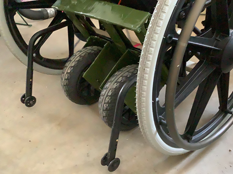 A cardboard frame covered in green tape with two motors sits in the lower back half of a wheelchair. This is the dimensioning prototype. 