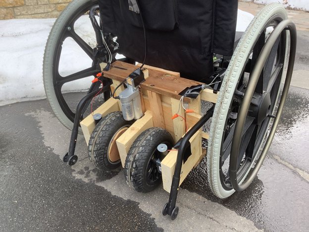 A wooden frame with two wheels sits on the back of a wheelchair positioned slightly below the seat. This was the propulsion prototype. 